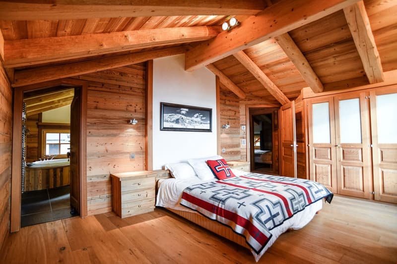 Rustic wooden cabin bedroom with vaulted ceiling, wooden walls and beams, large windows, a bed with a patterned blanket, and a framed artwork on the wall