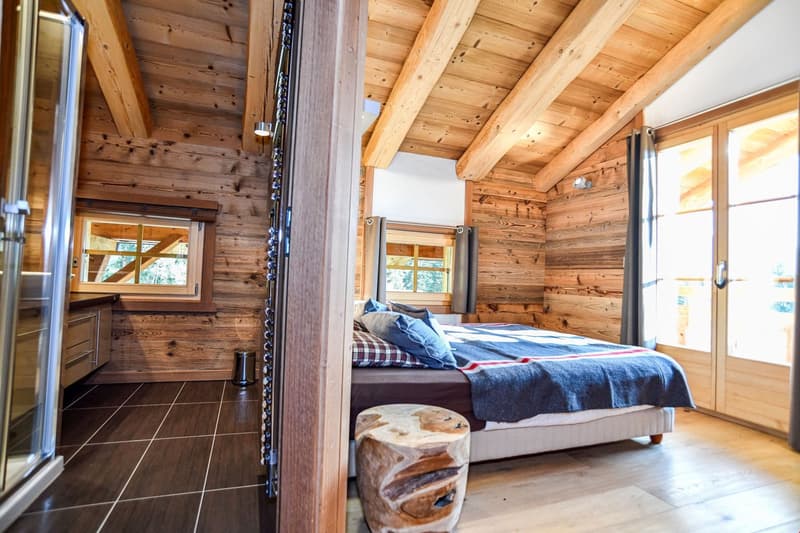 Cozy wooden cabin-style bedroom with large windows, wooden beams and paneling, a comfortable bed with plaid bedding, and a wooden stool or side table.