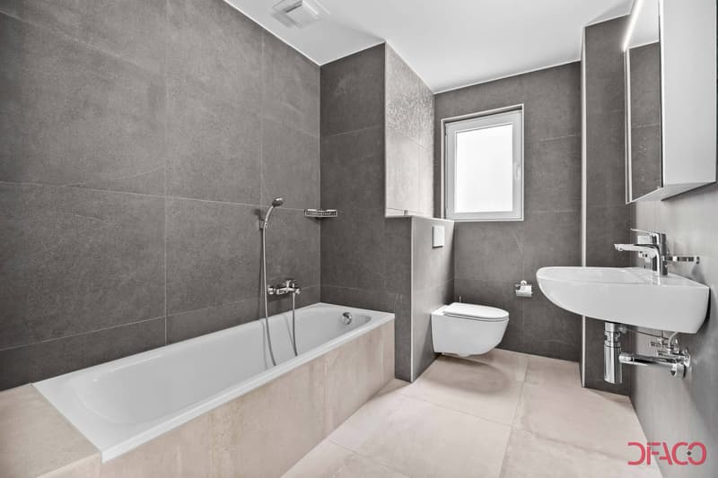 modern and minimalistic bathroom with gray tiles, white bathtub, white sink, white toilet, overhead shower, window