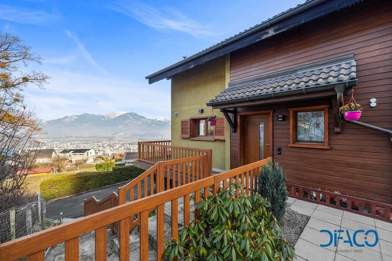 Exterior of a house with wooden exterior, balcony, windows with wooden shutters, surrounded by trees and bushes, mountain in the distance, wooden staircase, paved pathway.