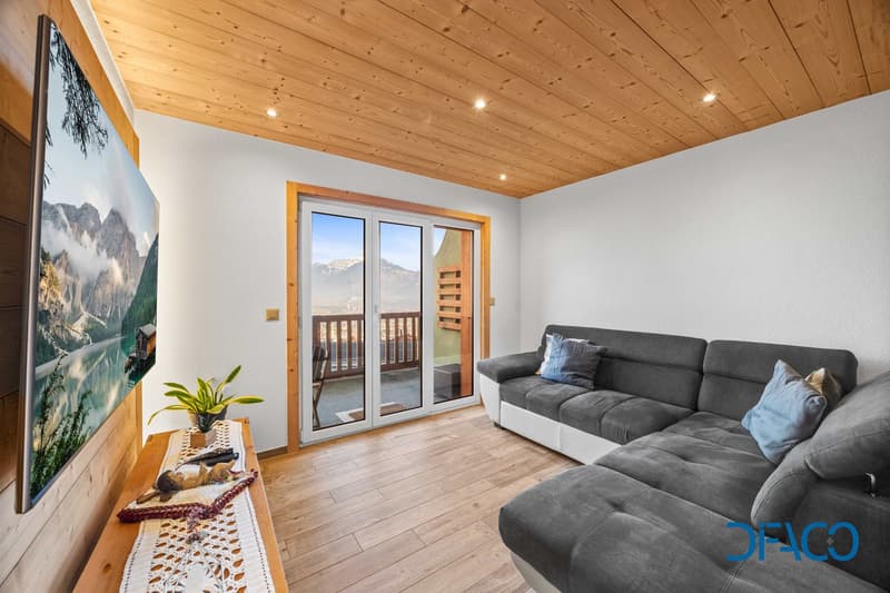 Living room with grey sofa, wooden floor, TV mounted on wall, sliding glass door leading to balcony, wooden ceiling, balcony with railings, view of mountains
