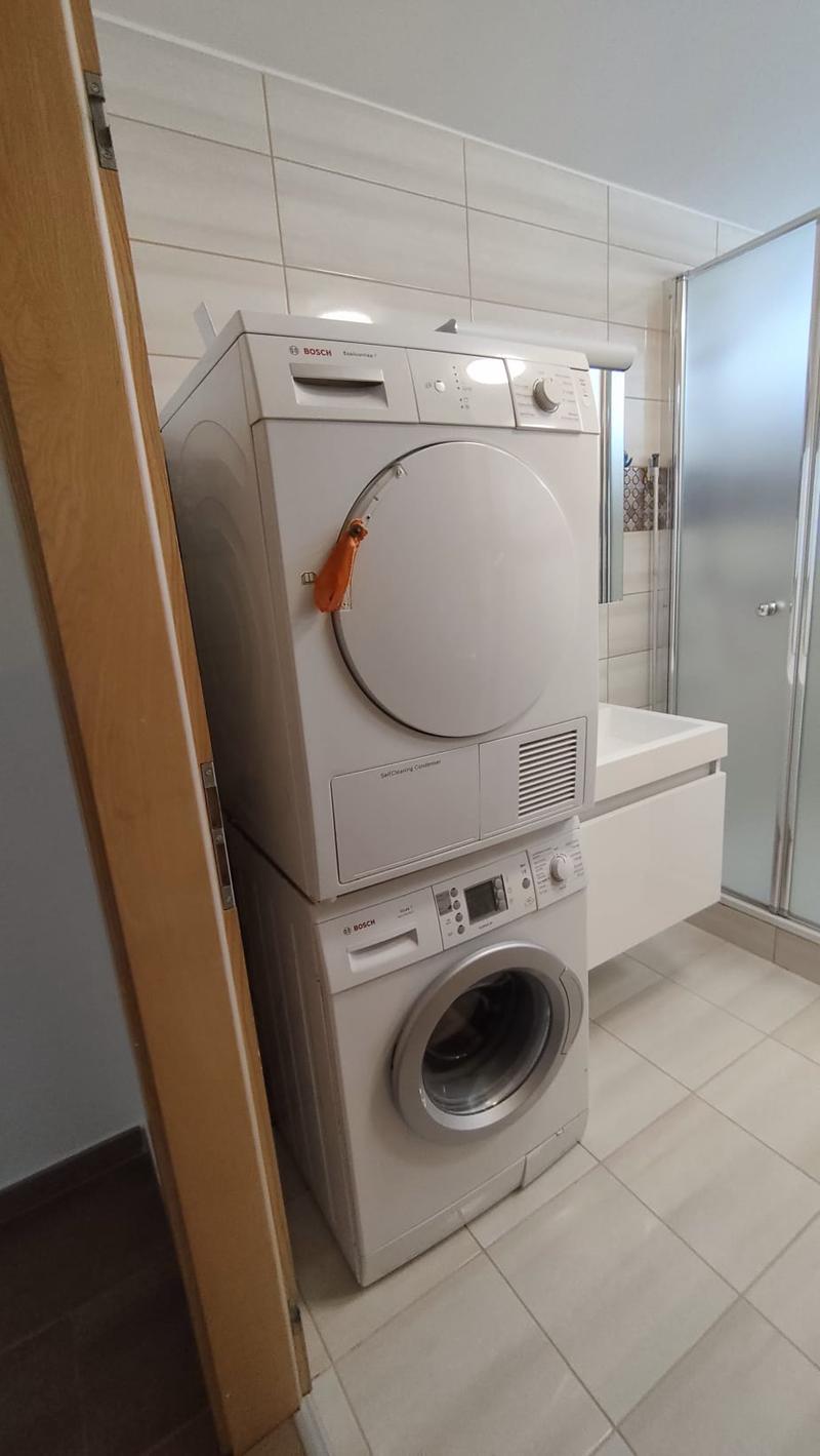 A bathroom with a white tiled floor, a white sink, a glass shower door, and a stack of white Bosch washing machines.