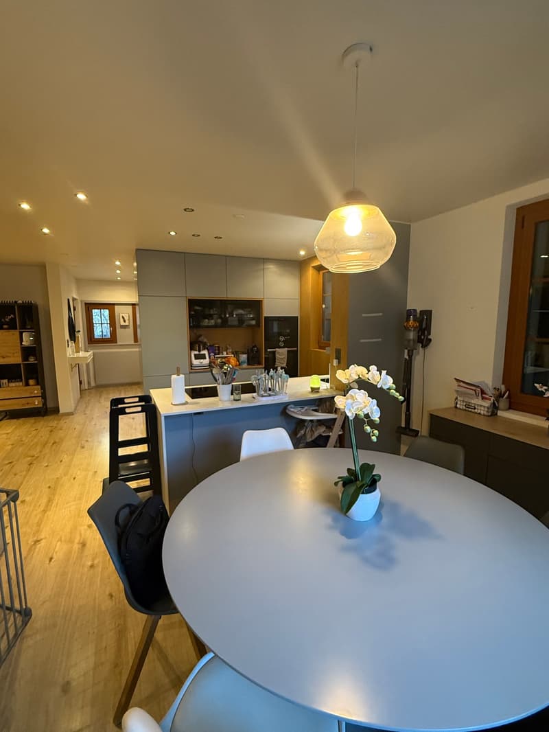 Modern kitchen-dining area with a round table, gray chairs, white flowers, wooden floor, gray cupboards, refrigerator, and a window.