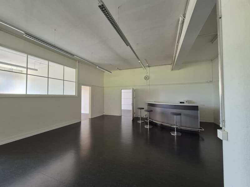 spacious room with white walls, black flooring, large glass windows, open door leading to another room, high ceiling with recessed lights, bar counter with stools