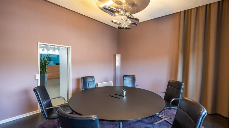 A meeting room with an oval table, black leather chairs, round chandelier, and a beige wall with curtains.