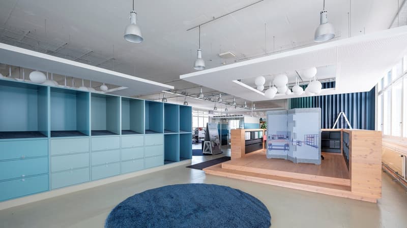 The interior of the office space features tall blue cabinets along one wall, modern office furniture, an elevated wooden platform with a gray backdrop, and pendant lamps on the ceiling.