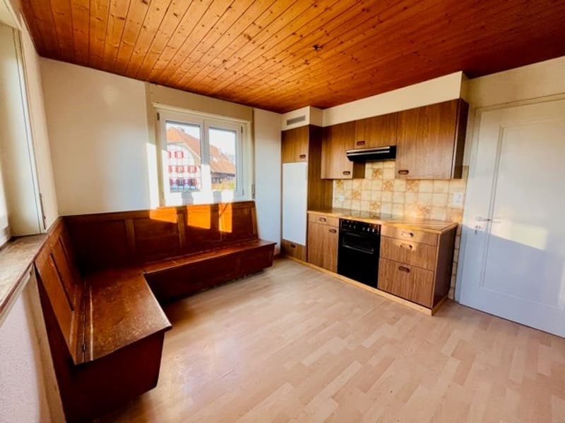 Empty room with wooden floor, wooden bench, wooden cabinets, white walls, wooden ceiling, window, black oven, and white refrigerator