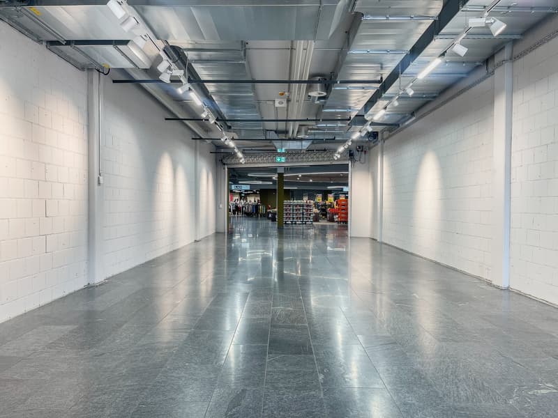 Empty hallway with white walls, gray tiled floor, and overhead lighting fixtures.