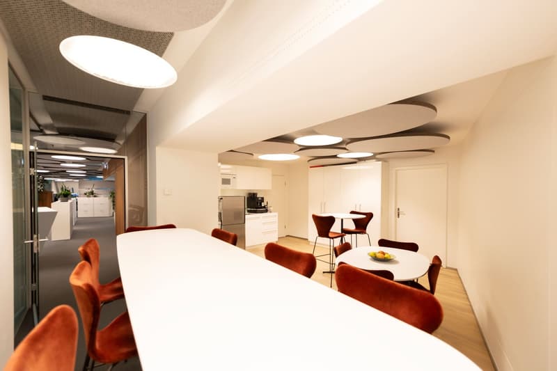 Open-concept kitchen and dining area with a large white table, red upholstered chairs, and a modern lighting fixture with circular panels. The kitchen area has white cabinets and a stainless steel appliance, likely a dishwasher.
