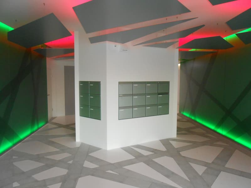 The image shows a modern office space with a white wall and a set of lockers or storage units. The ceiling has geometric shapes and patterns with colored lighting, creating a visually striking and dynamic atmosphere. The floor has a geometric tile pattern