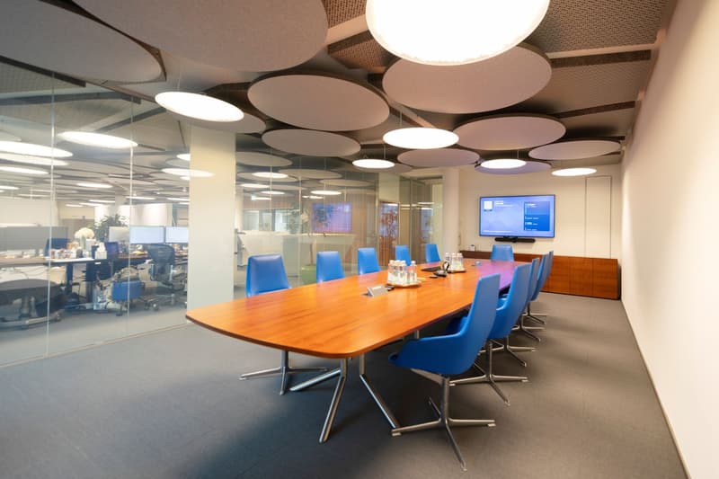 Large wooden conference table with blue chairs, modern lighting fixtures on the ceiling, glass walls separating the conference room from the open office space, and a large display screen on the wall.
