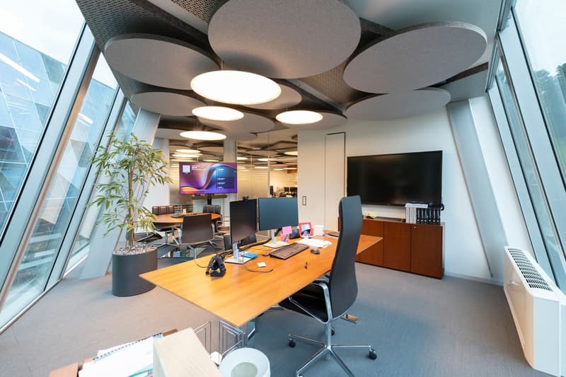 Modern office space with large windows, circular lighting fixtures, computer workstations, and a TV display on the wall