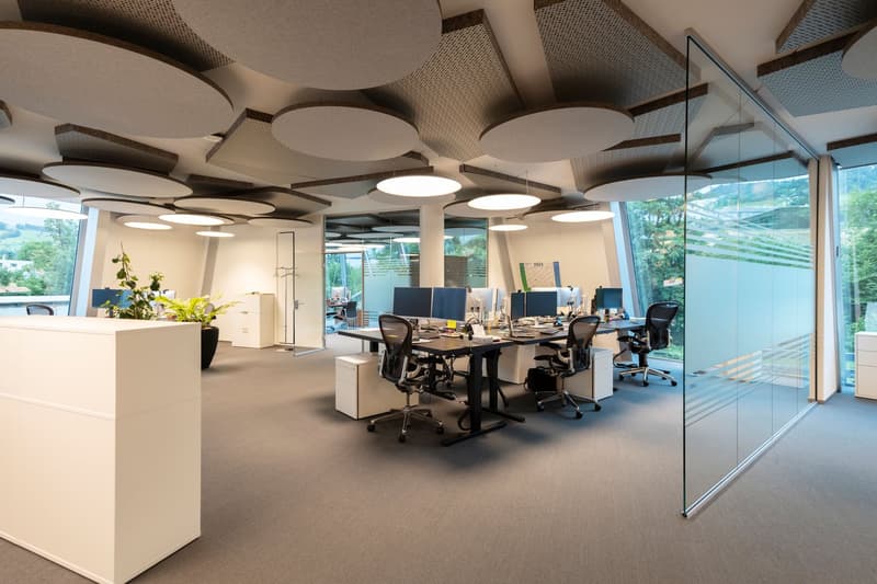 Open-plan office space with modern furniture and lighting, glass walls, and a view of the outdoors