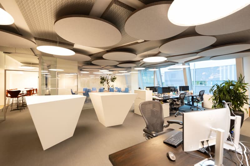Open-plan office space with modern, minimalist design. Large circular light fixtures on the ceiling, white desks and chairs, computers and monitors, and potted plants. Glass walls and windows provide natural light.
