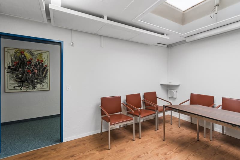A rectangular room with white walls, wooden floor, a desk, chairs and a painting.