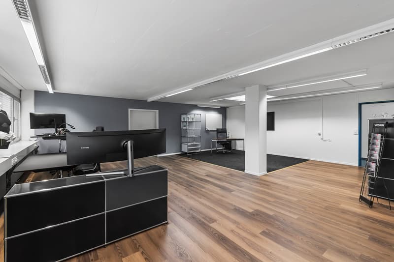 Spacious office room with wooden floor, white ceiling, monitors, desk, chairs, shelf, and storage unit.