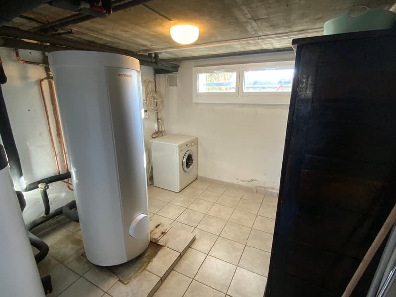 Basement room, white walls, tiled floor, white washing machine, tall white water heater, window, storage cabinet