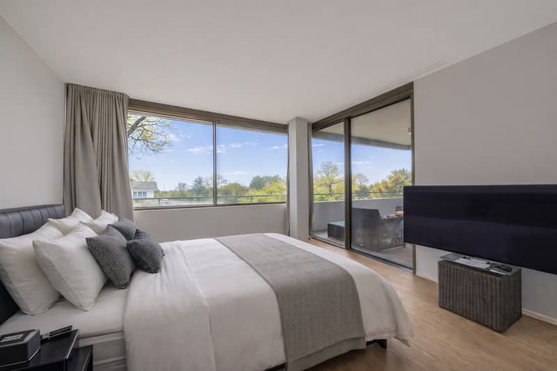 Large bed, white sheets and grey blanket, sliding windows with balcony, TV on stand, wooden floor, light walls
