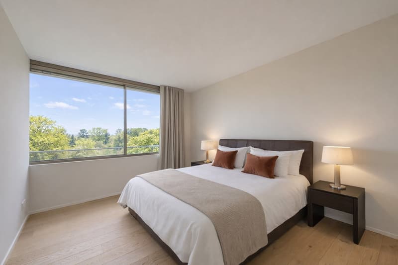 Bed with white bedding, two nightstands with lamps, large window with curtains, hardwood floor, no visible appliances