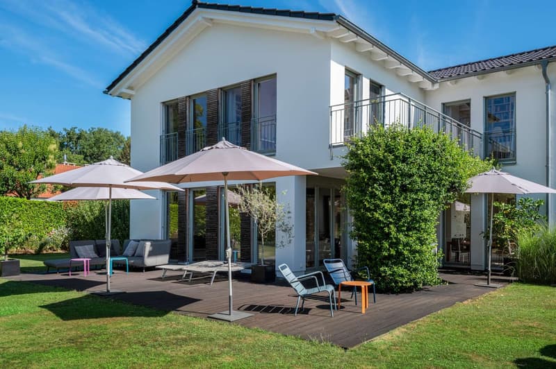Two-story white house with a balcony, surrounded by a well-maintained garden. The house has large windows and a patio area with outdoor furniture and umbrellas, providing a comfortable outdoor living space.