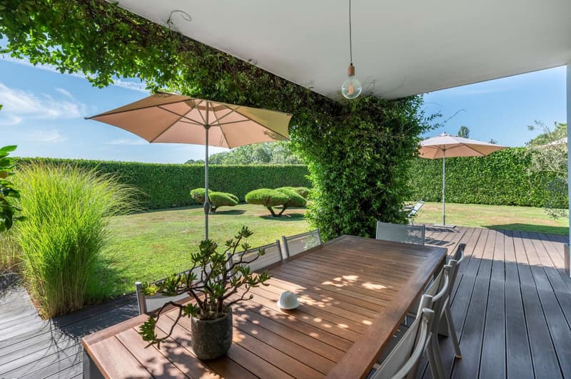 Wooden deck with dining table and chairs, large patio umbrellas, lush greenery and landscaping, including hedges and potted plants