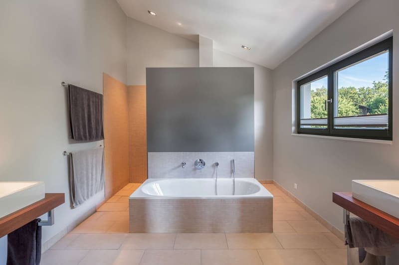 Large bathroom with a freestanding bathtub, gray and white tile flooring, gray accent wall, and a window overlooking the outdoor area.