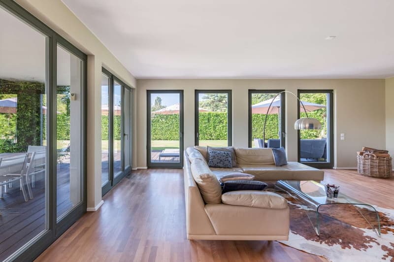 Spacious living room with large windows overlooking a garden, hardwood floors, beige sofa, coffee table, and a floor lamp