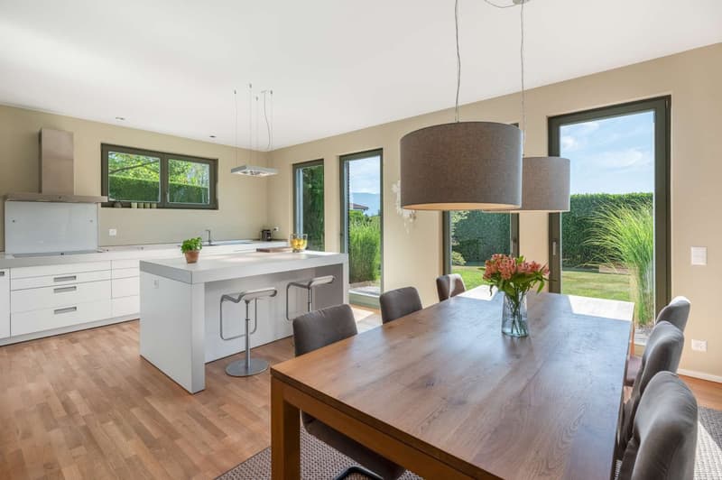 Open-concept kitchen and dining area with large windows overlooking a garden, hardwood floors, modern kitchen with white cabinets and island, wooden dining table with chairs, pendant lighting fixtures