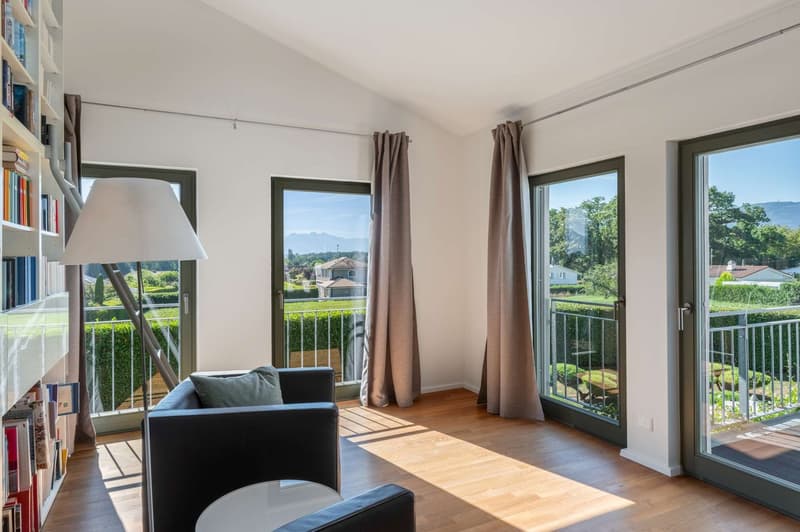 Spacious living room with large windows, hardwood floors, black sofa, bookshelf, and access to a balcony or terrace overlooking a scenic outdoor view