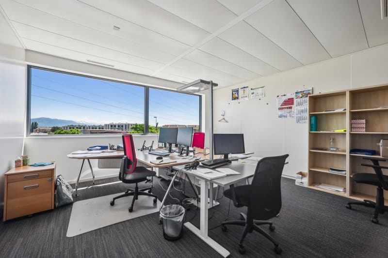 Spacious office with large windows providing a view of the city, multiple desks and computer workstations, shelving units, and office chairs.