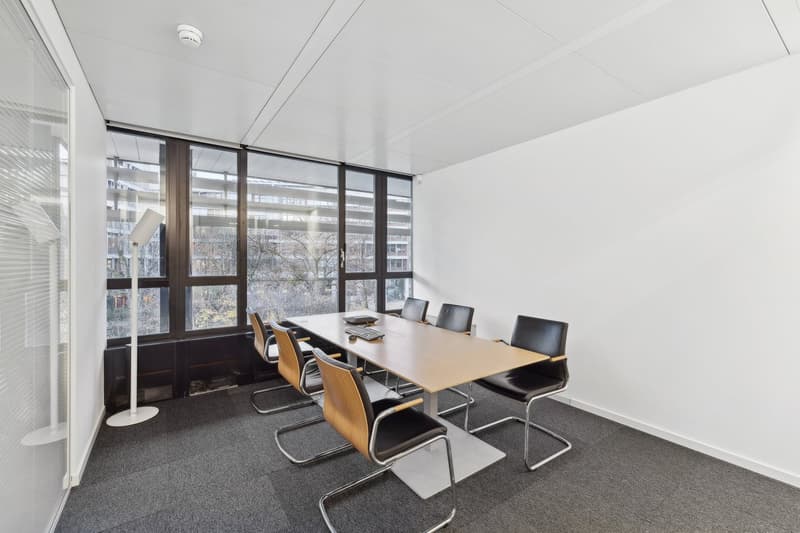 A rectangular meeting room with large windows, gray carpet, wooden desk, and modern office chairs.