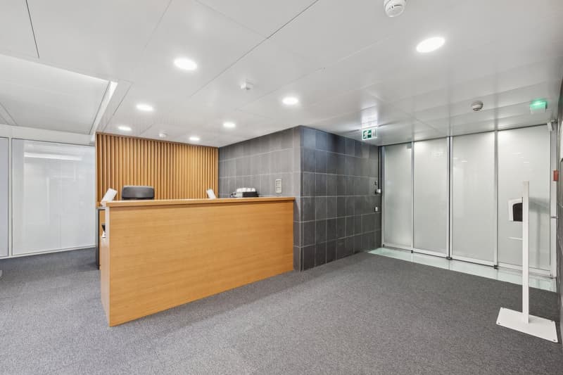 A rectangular reception area with a wooden counter, a black office chair, and gray flooring. To the left, there's a white office chair and a desk with an object. On the right, there's a gray wall with glass doors.