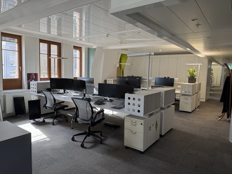 Open office space with white cubicles, computer stations, glass and wooden windows, radiators, central pathway, speakers, and indoor plants.