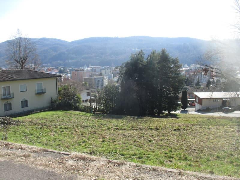 The image shows a scenic view of a city surrounded by mountains. There are residential buildings with balconies visible, as well as trees and greenery in the foreground. The city skyline can be seen in the distance, with tall buildings and a hazy, mountai