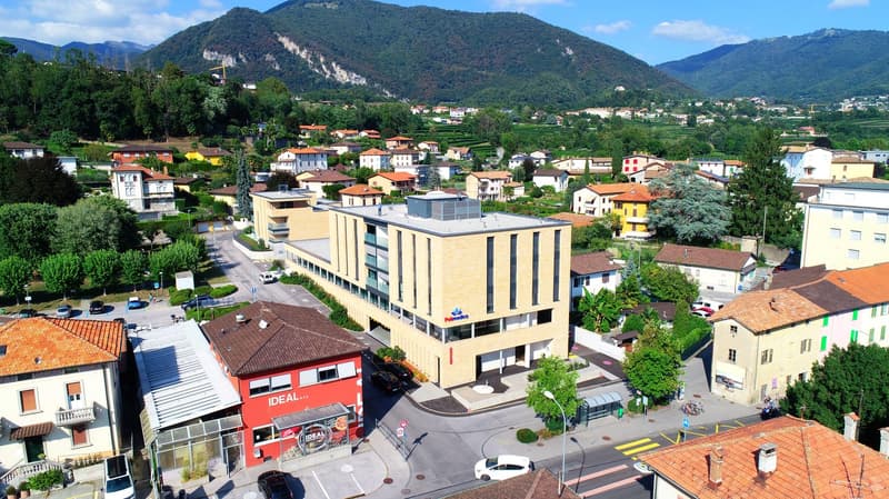 Aerial view of a town, with numerous buildings, red roofs, surrounded by green vegetation, mountains in the background.
