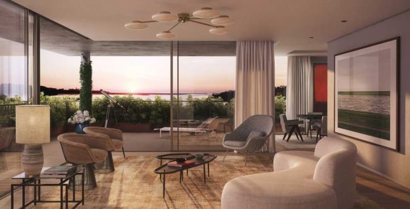 Spacious living room with large windows offering a scenic view of the outdoors, including a balcony or terrace. The room features comfortable seating, a dining area, and decorative elements like artwork and lighting fixtures.