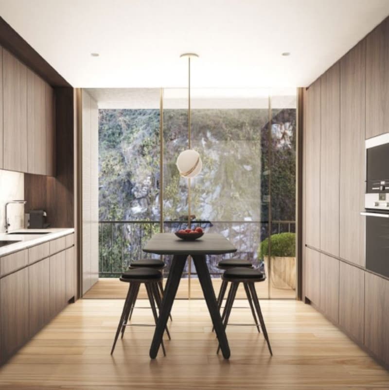 The image shows a modern kitchen and dining area with large windows overlooking a scenic outdoor view. The kitchen features wood-paneled cabinets and a minimalist design. The dining table is a simple, dark-colored wooden table with black stools. The room