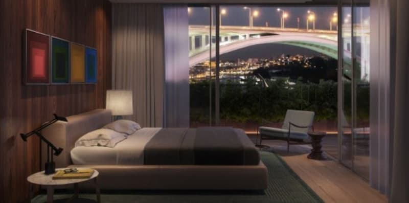 Spacious bedroom with large windows overlooking a night cityscape, wooden walls, modern furniture including a bed, armchair, and side table with a lamp