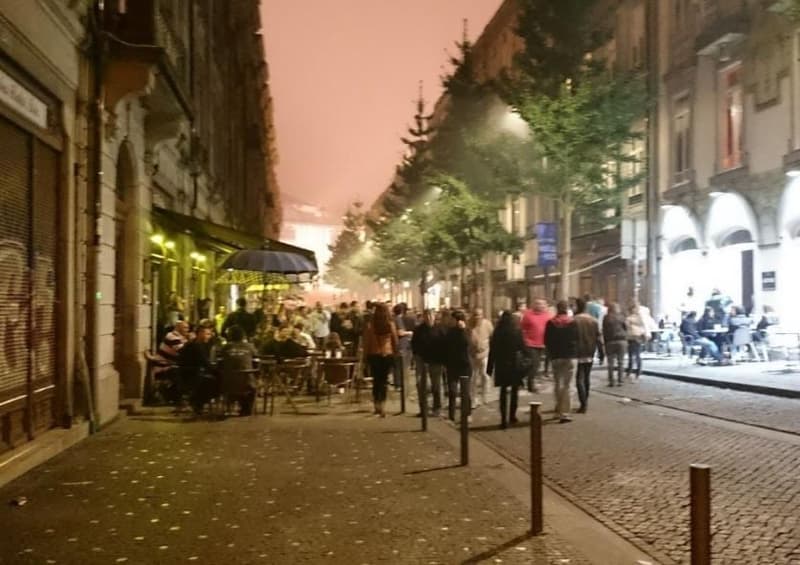 Nighttime street scene with people dining at outdoor cafes, trees lining the street, and buildings with balconies along the street