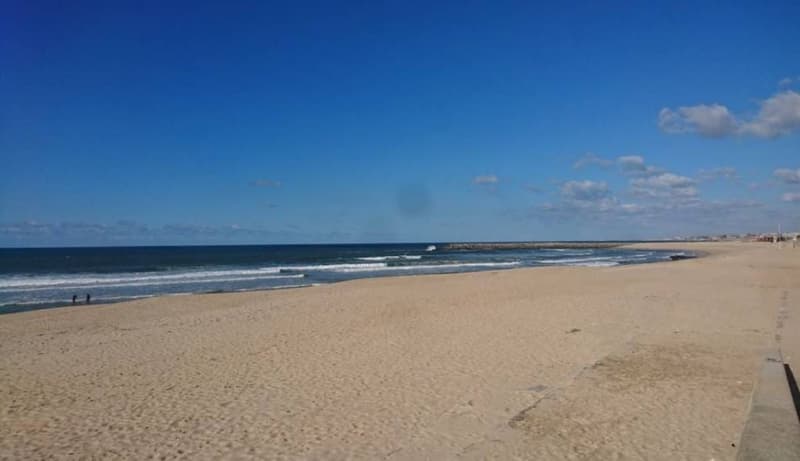 The image shows a wide, sandy beach with waves crashing along the shoreline. The sky is a deep blue color with some clouds, and the horizon line is visible in the distance. There are a few people walking along the beach.