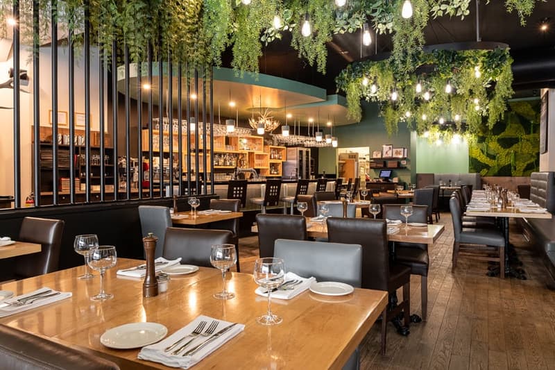 Upscale restaurant with a cozy and modern interior design. The space features wooden tables, leather chairs, and a bar area with a large selection of wines and liquors. The ceiling is adorned with hanging greenery and string lights, creating a warm and in