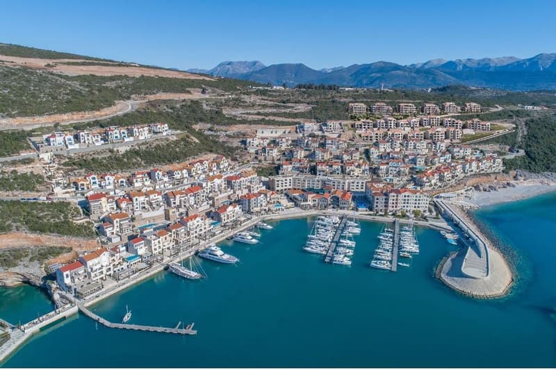 coastal town with buildings and a harbor, boats docked, mountain range in the background