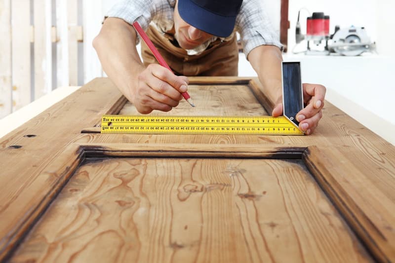 Person wearing blue cap and tan apron using a yellow measuring tape with a pencil to draw on a wooden surface in a workshop setting with tools in the background.