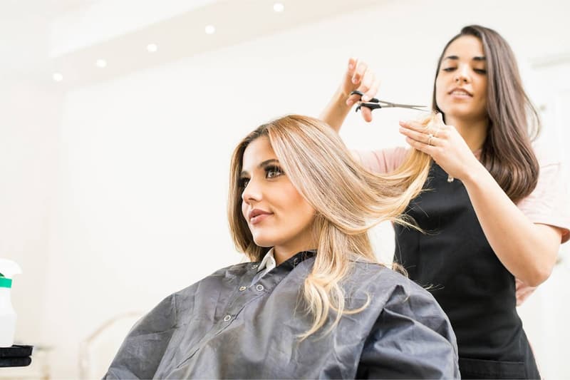 A woman with long blonde hair is getting her hair styled by another woman in a hair salon. The stylist is using scissors to cut and shape the client's hair.
