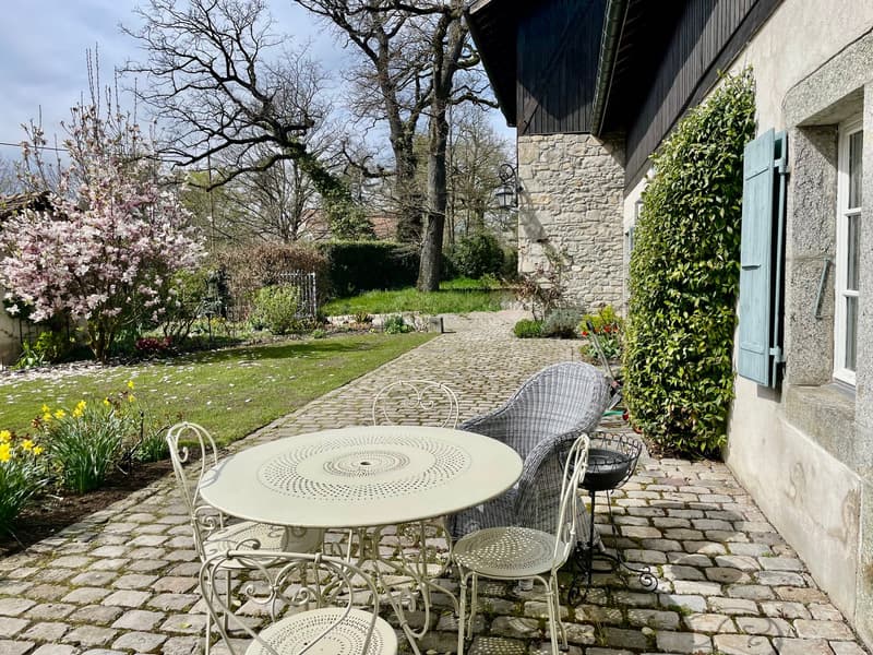 Two-story stone house with a cobblestone path leading to a patio area with wrought iron furniture, surrounded by lush greenery, flowering trees, and a well-maintained garden.