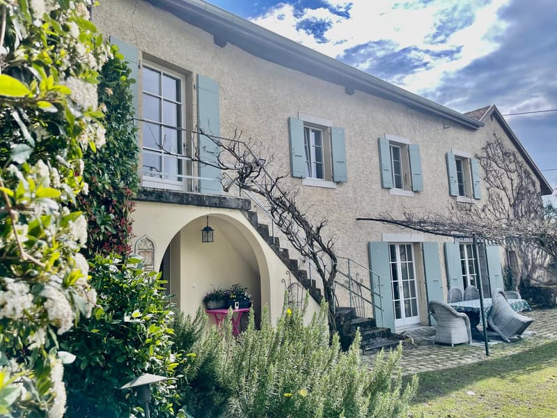 Two-story stone house with arched entryway, green shutters, and a balcony. The house is surrounded by lush greenery and a paved patio area with outdoor furniture.