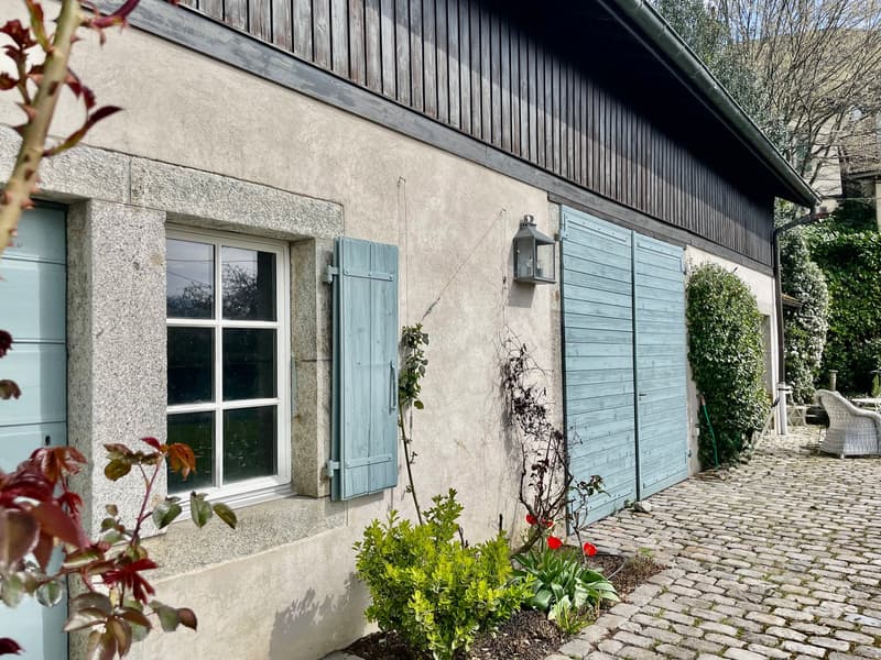 2-story wooden house with stone exterior, blue shutters, large windows, cobblestone path, and landscaped garden with plants and flowers