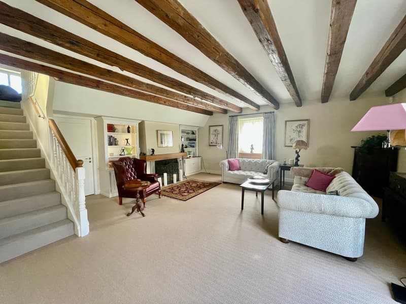 Spacious living room with exposed wooden beams on the ceiling, white walls, hardwood floors, a large L-shaped sofa, an armchair, a coffee table, and a fireplace. The room also has built-in shelves and a staircase leading to another level.