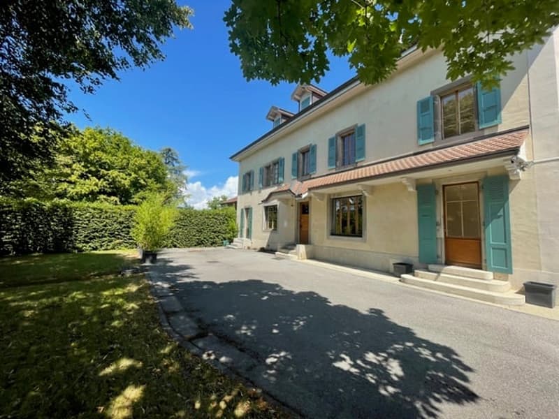2-story house with beige exterior, green shutters, large windows, and a paved driveway or parking area. The house is surrounded by lush greenery and trees.