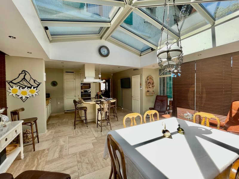 Dining room with wooden tiles, glass roof, chandelier, clock, table, chairs, bar stools, counter, kitchen, fireplace, wall decorations, shelves, microwave